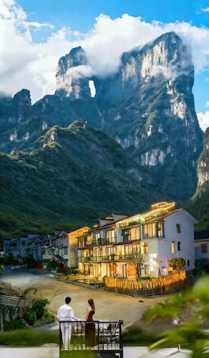 Un hotel moderno con fachada de vidrio, situado en una zona montañosa con un pico rocoso destacado al fondo.