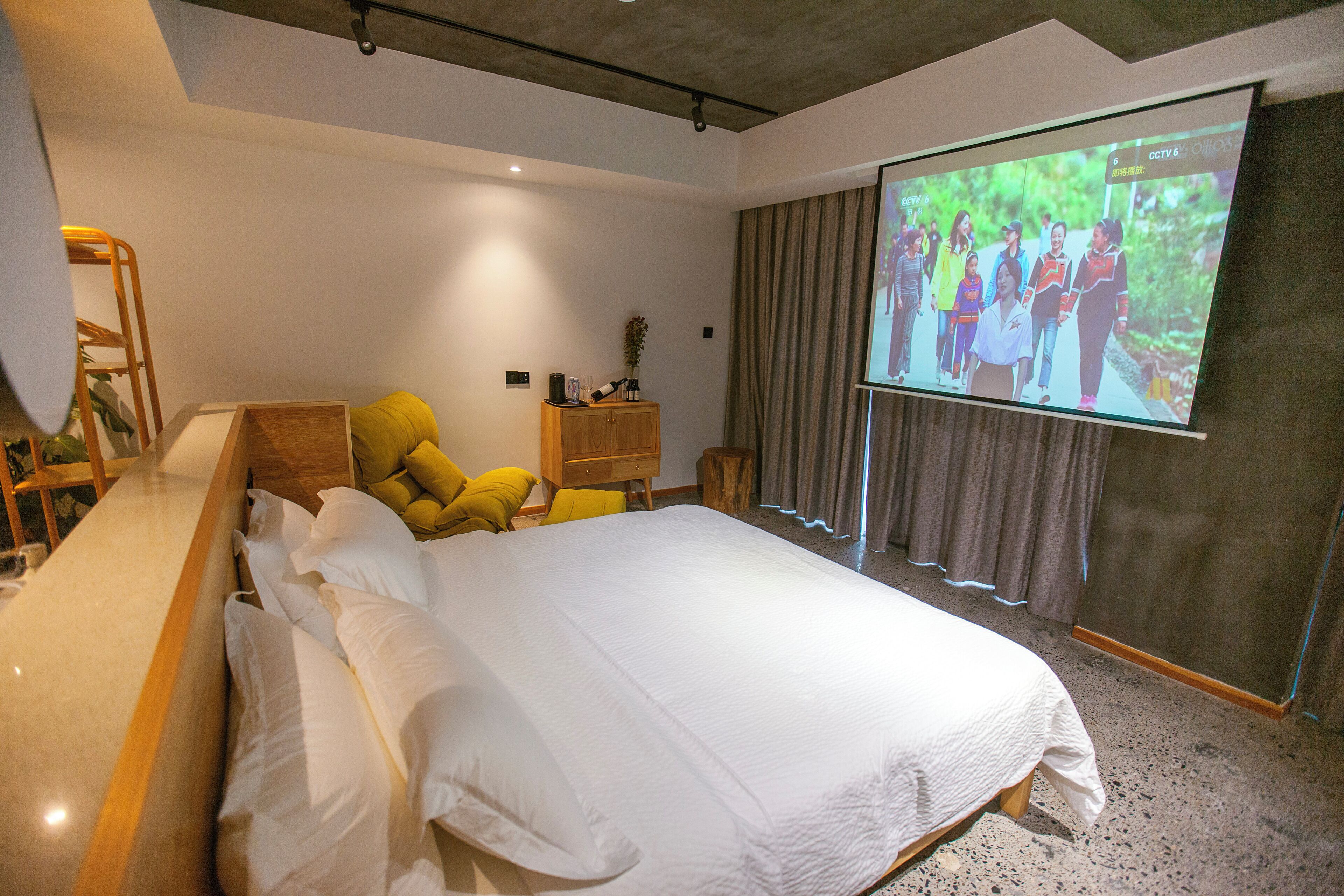Una habitación de hotel moderna con una cama grande, una mesita de noche de madera, un sillón amarillo y una pantalla de proyector mostrando un grupo de personas.