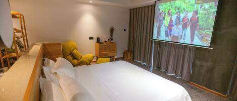 Una habitación de hotel moderna con una cama grande, una mesita de noche de madera, un sillón amarillo y una pantalla de proyector mostrando un grupo de personas.