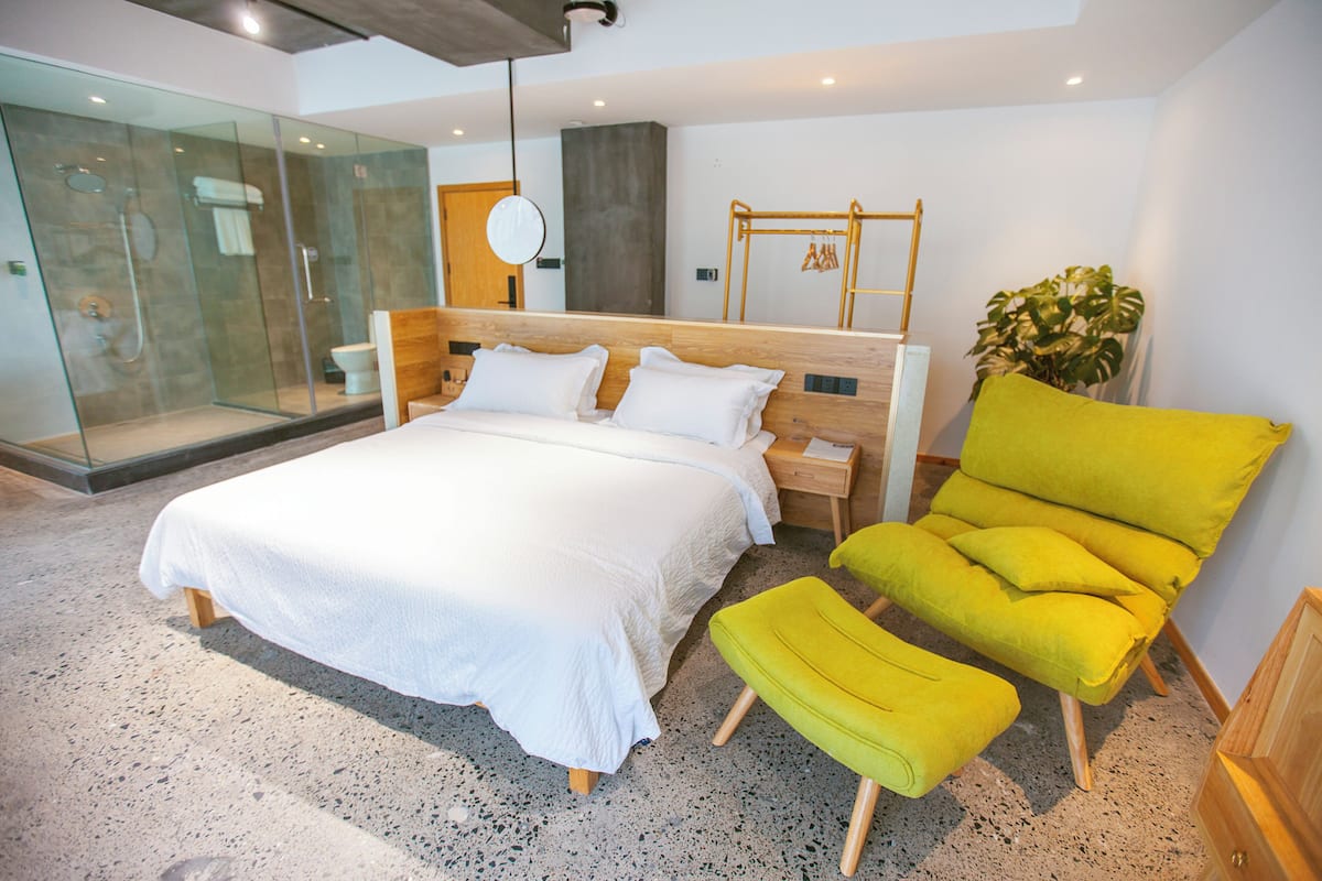 A modern bedroom with a large bed, a glass shower enclosure, a wooden headboard, a yellow armchair, and a small plant.