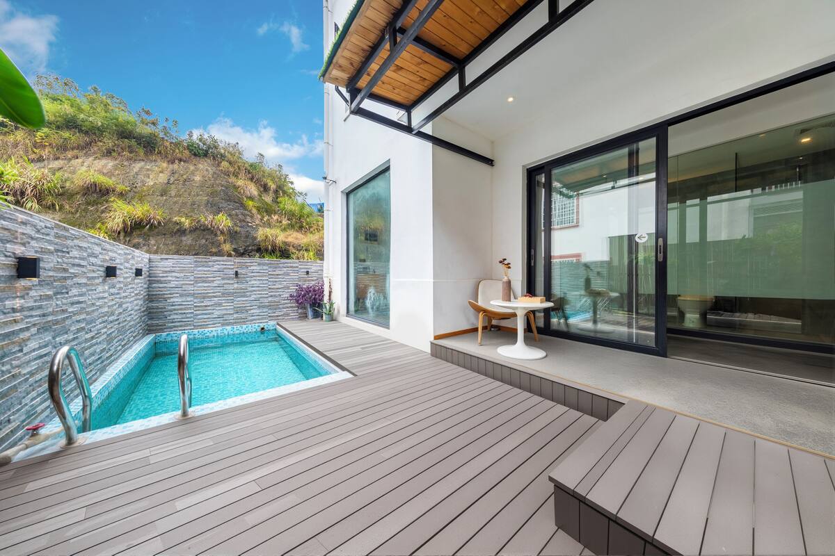 A modern outdoor pool area with a wooden deck, a seating area with a table and chairs, and a glass door leading to a covered patio.