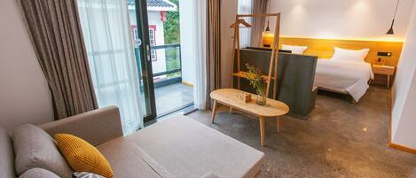 A hotel room with a sofa, a bed, a wooden nightstand, a hanging coat rack, and a view of the outside.