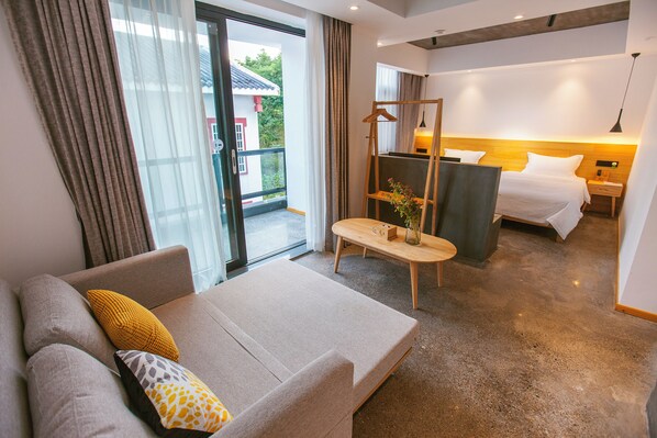 A hotel room with a sofa, a bed, a wooden nightstand, a hanging coat rack, and a view of the outside.