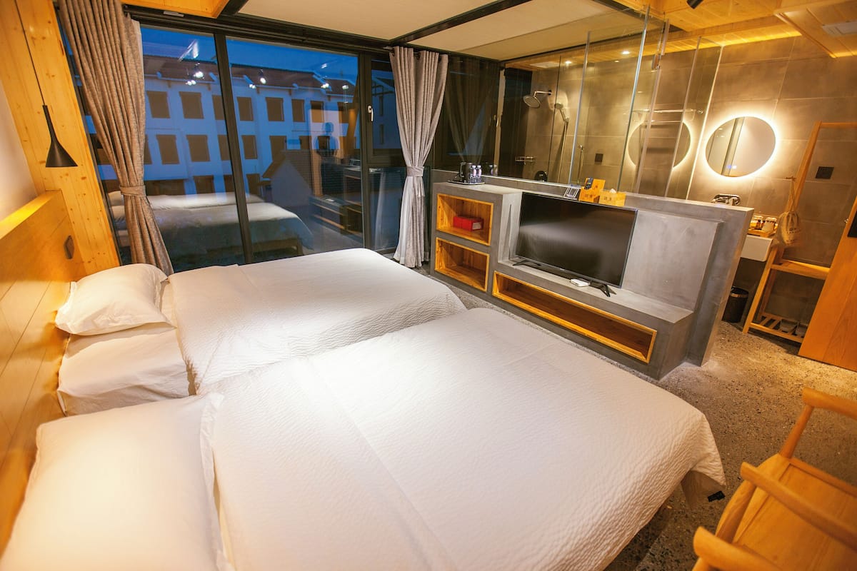 A modern hotel room with two beds, a TV, a mirror, and a view of the city at dusk.