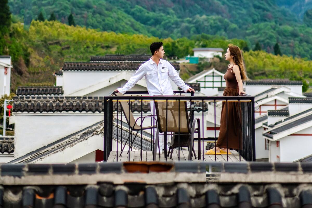 A couple on a rooftop terrace with traditional Chinese architecture, overlooking a mountainous landscape with clouds.
