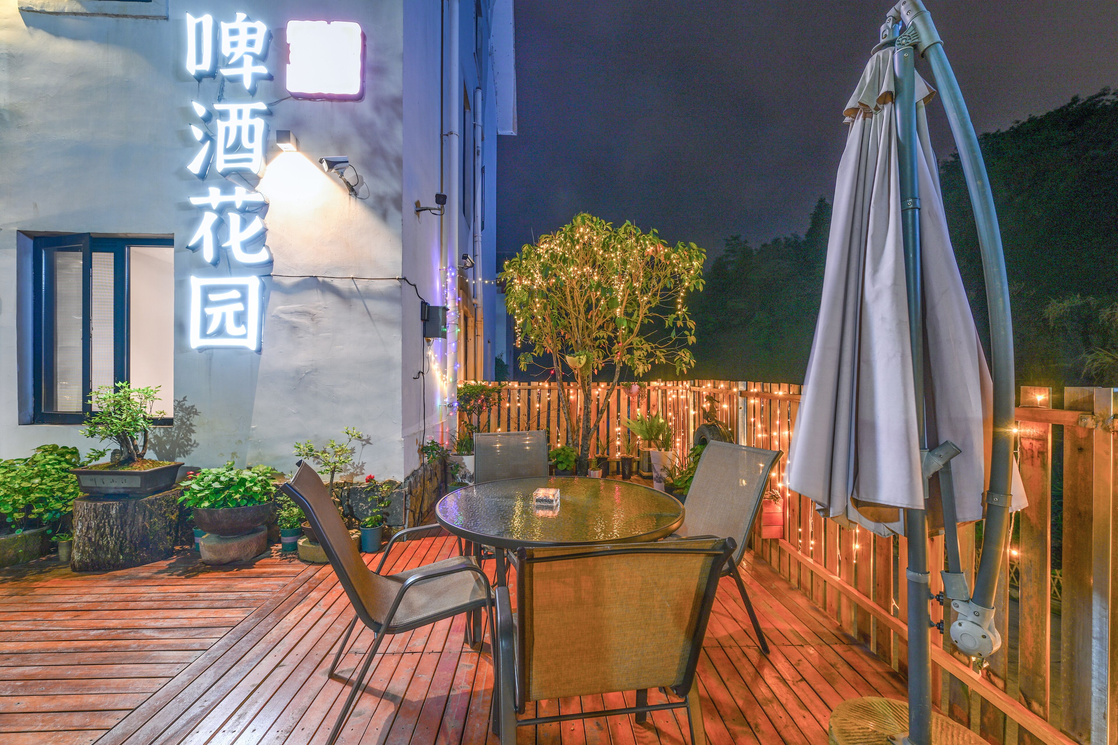 Una terraza de madera con una mesa redonda y sillas, rodeada de macetas con plantas y una valla de madera con luces colgantes.