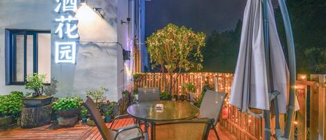 Una terraza de madera con una mesa redonda y sillas, rodeada de macetas con plantas y una valla de madera con luces colgantes.