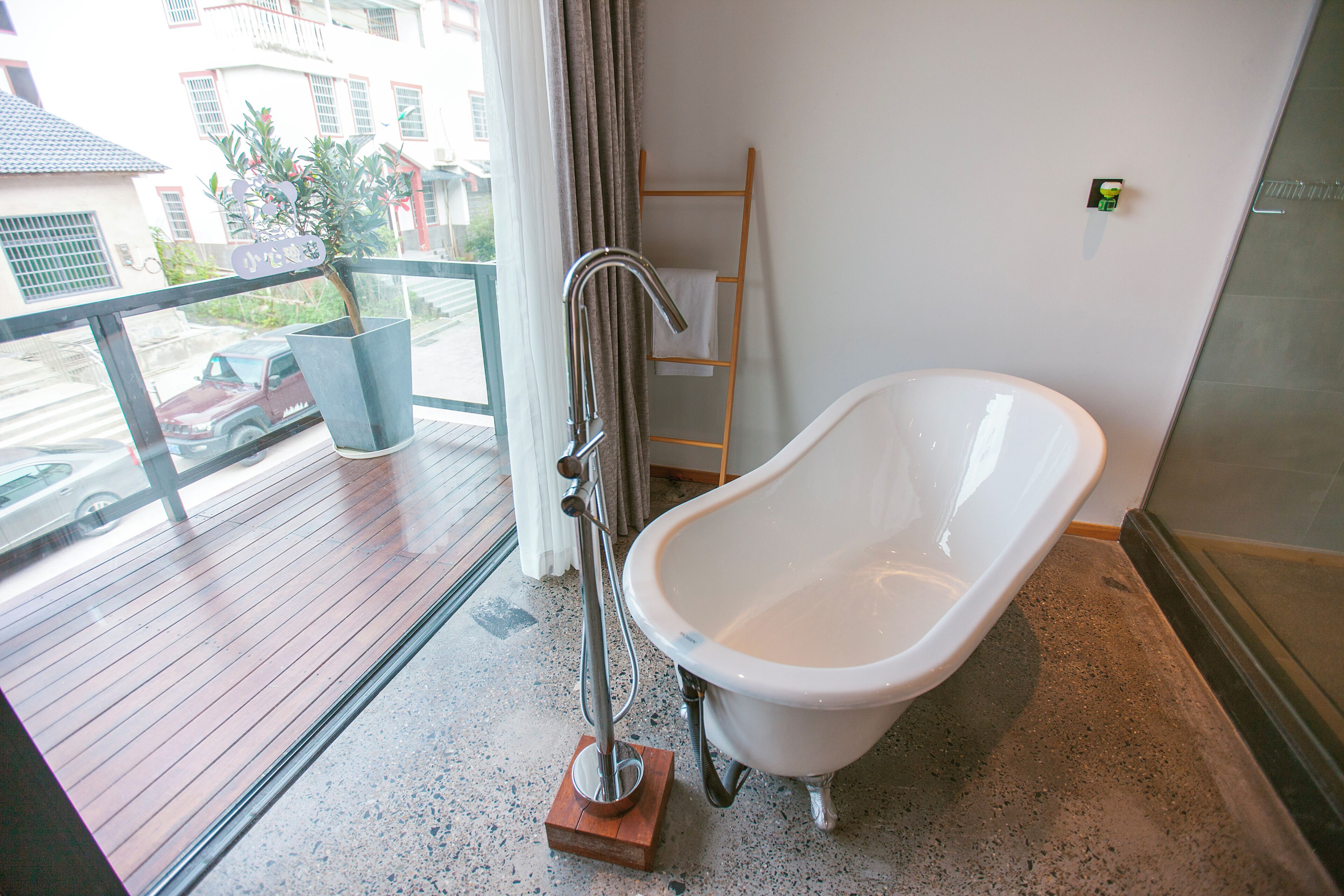 A modern bathroom with a freestanding bathtub, a wooden drying rack, and a view of the street through a large window.