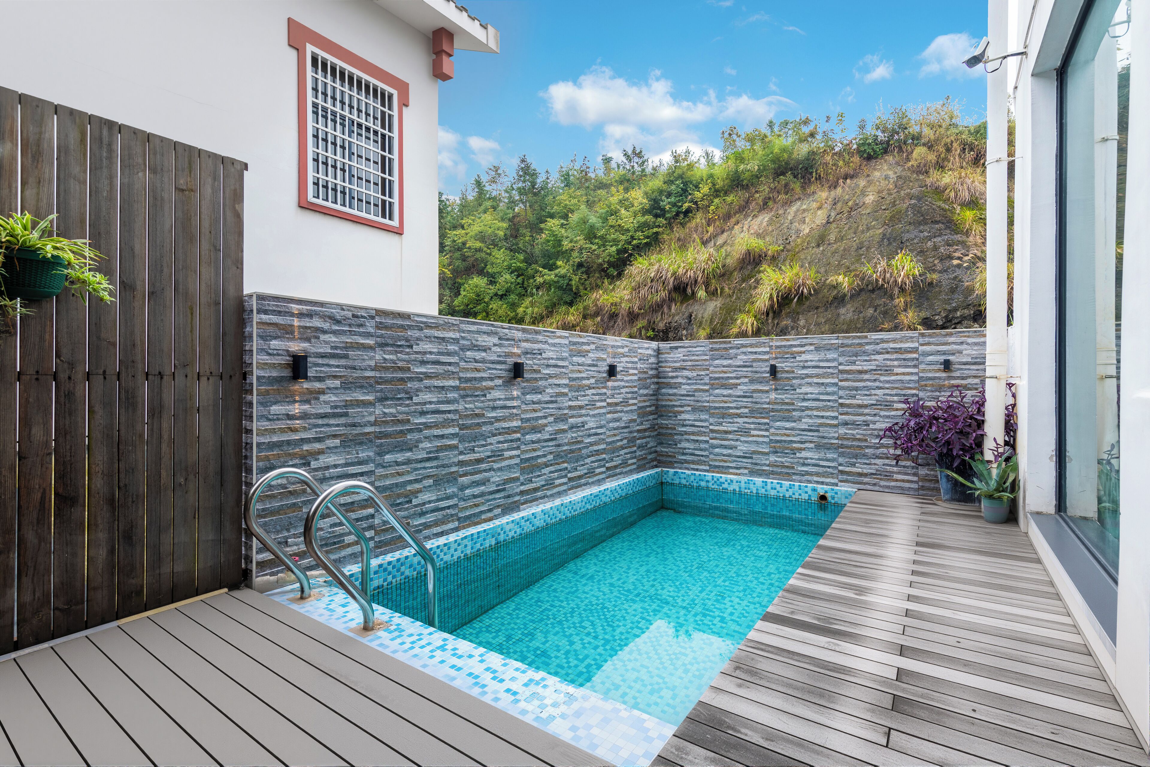 A swimming pool with a wooden deck, stone wall, and a window with a red frame.