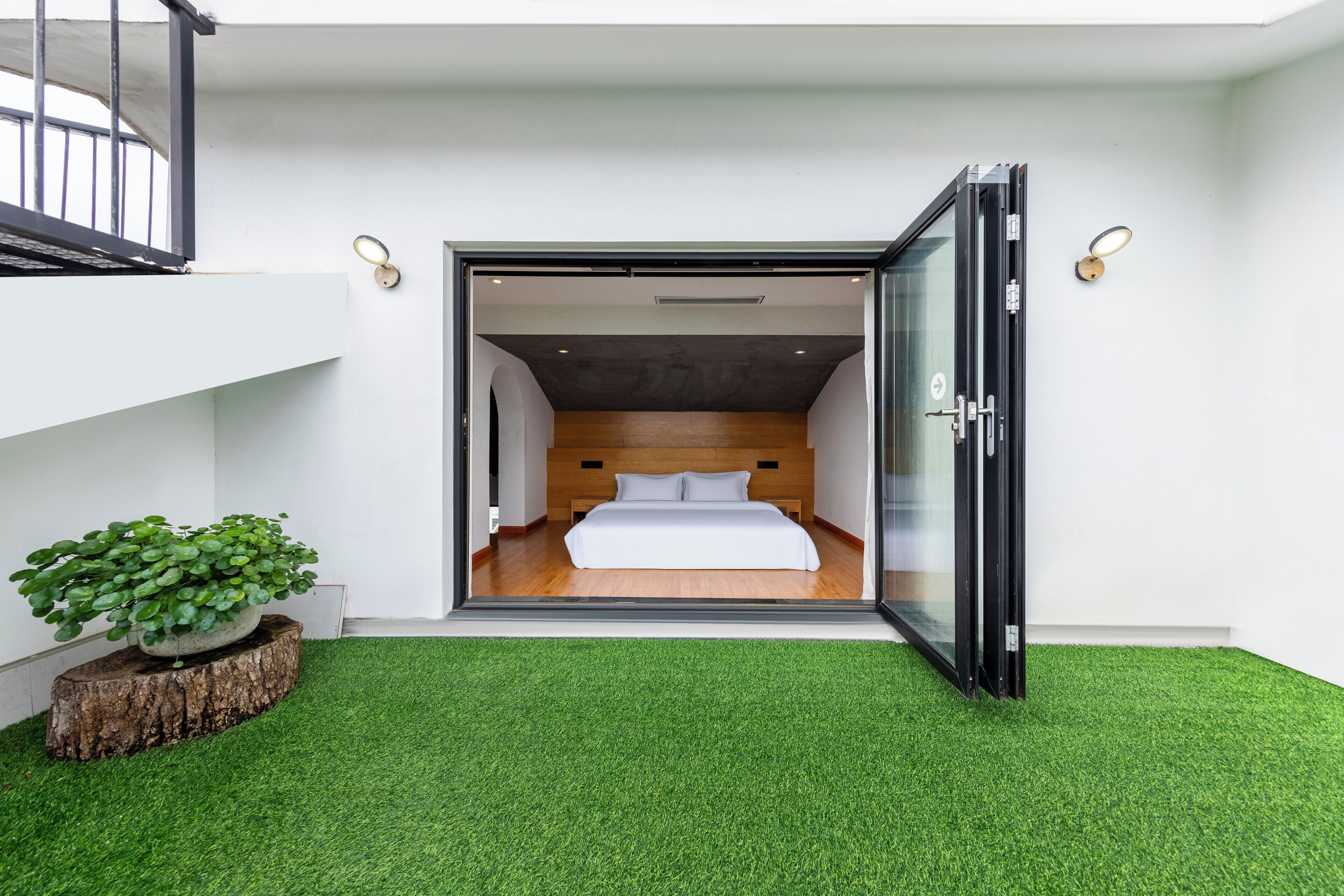 A modern bedroom with a glass door leading to a balcony, a wooden headboard, and a potted plant.