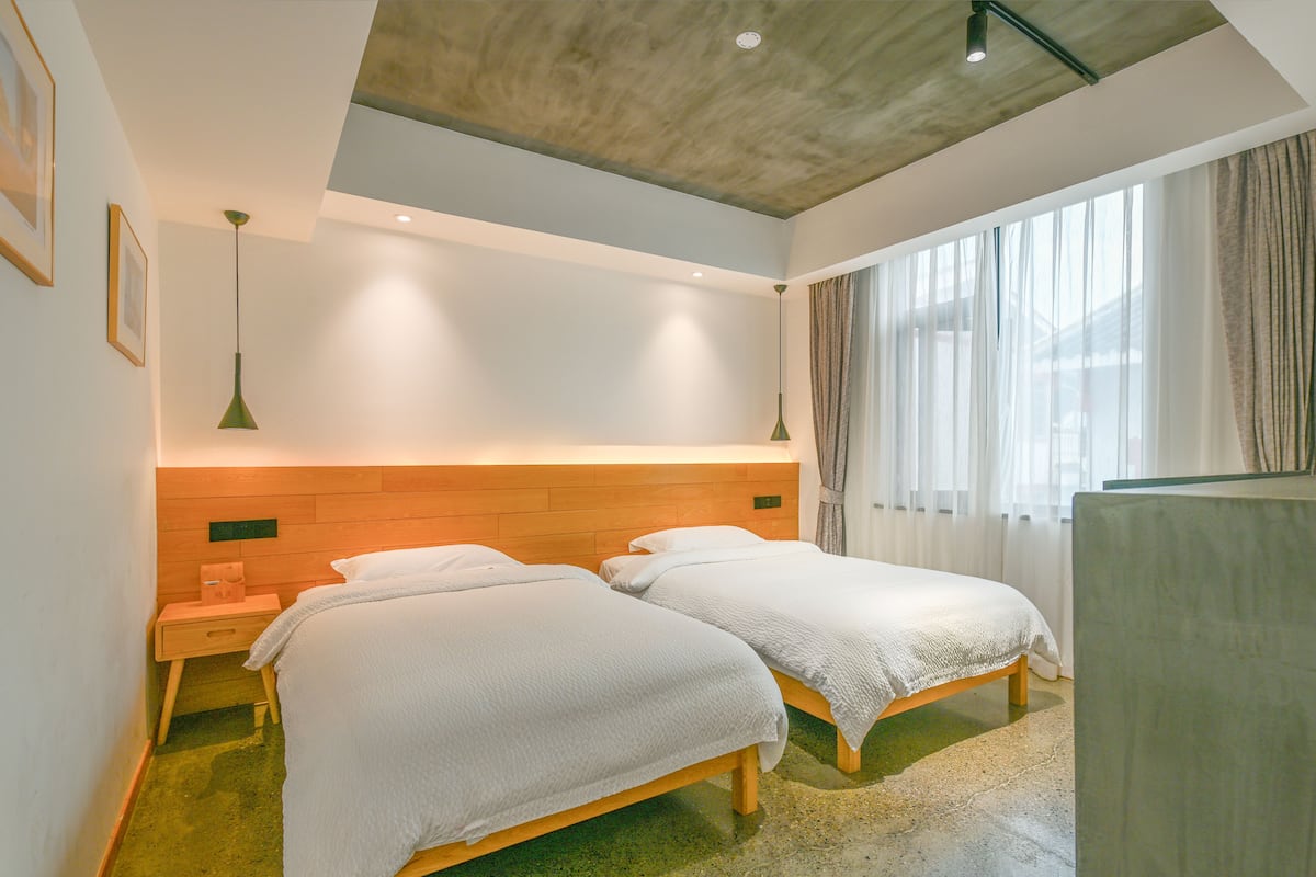 A hotel room with two beds, a wooden headboard, a small bedside table, and a large window with curtains.