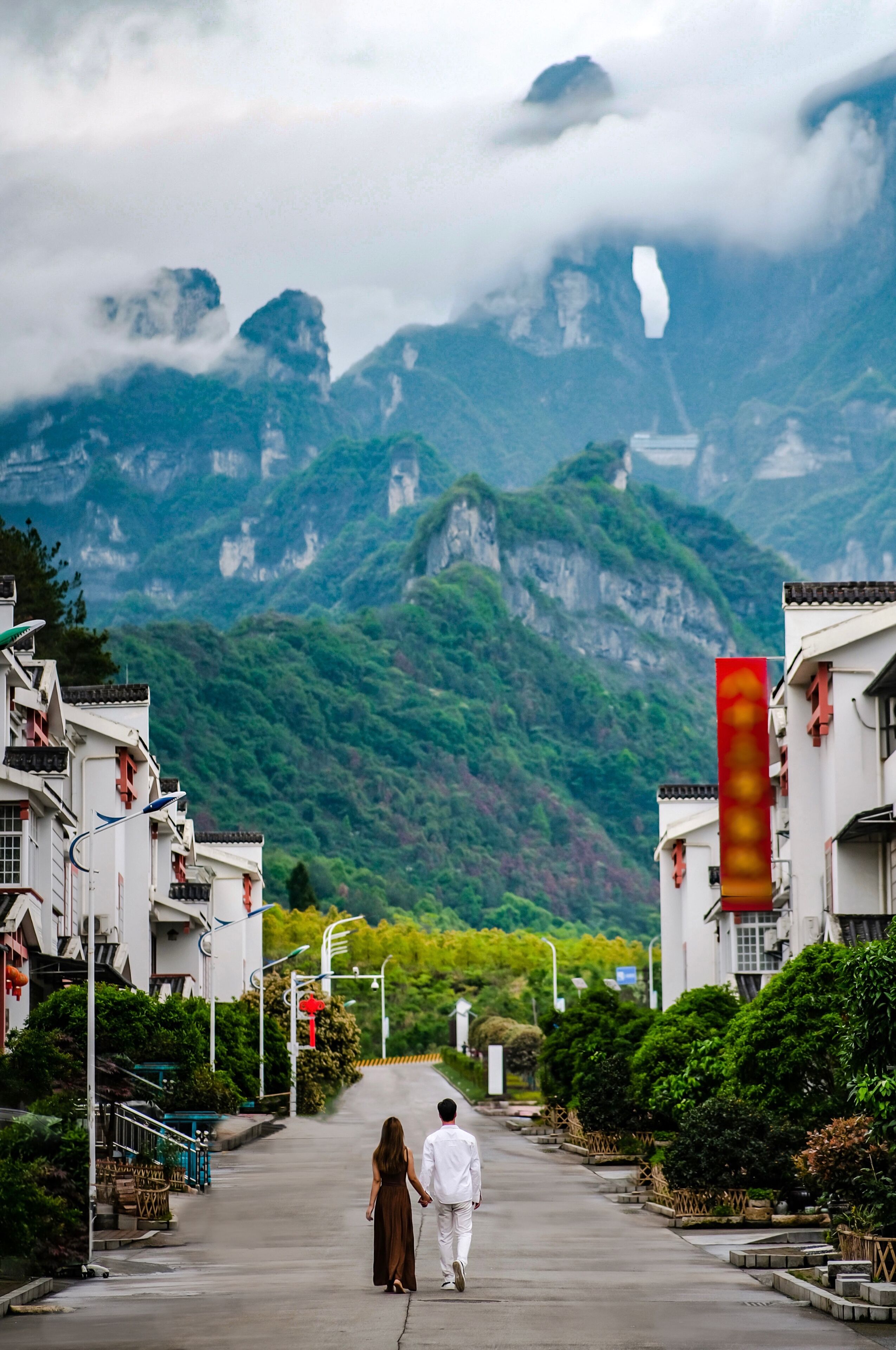 A couple walking down a street lined with traditional buildings, leading towards a mountainous landscape.