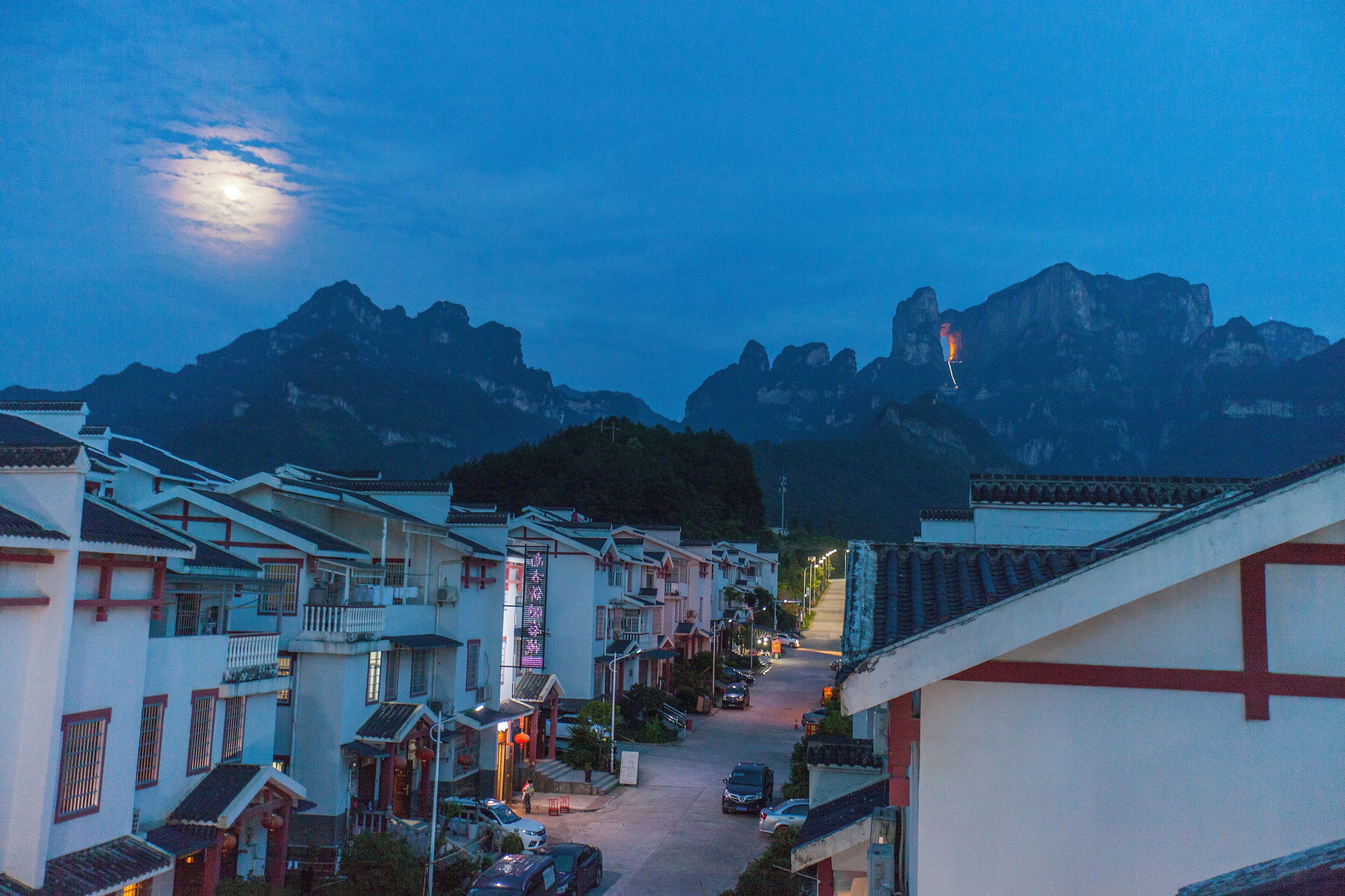 Uma rua tradicional numa vila, com edifícios, céu claro e montanhas ao fundo.