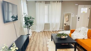 En moderne stue med trægulv, et sort sofabord med en plante, en orange sofa og en hvid, blød m åtte.
