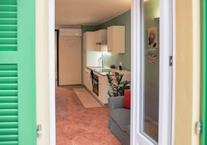 A compact kitchen with white cabinets, a sink, and a countertop. There is a grey sofa and a plant in the living area.