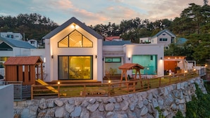 A modern house with a large glass entrance, a stone retaining wall, and a wooden deck with lights.