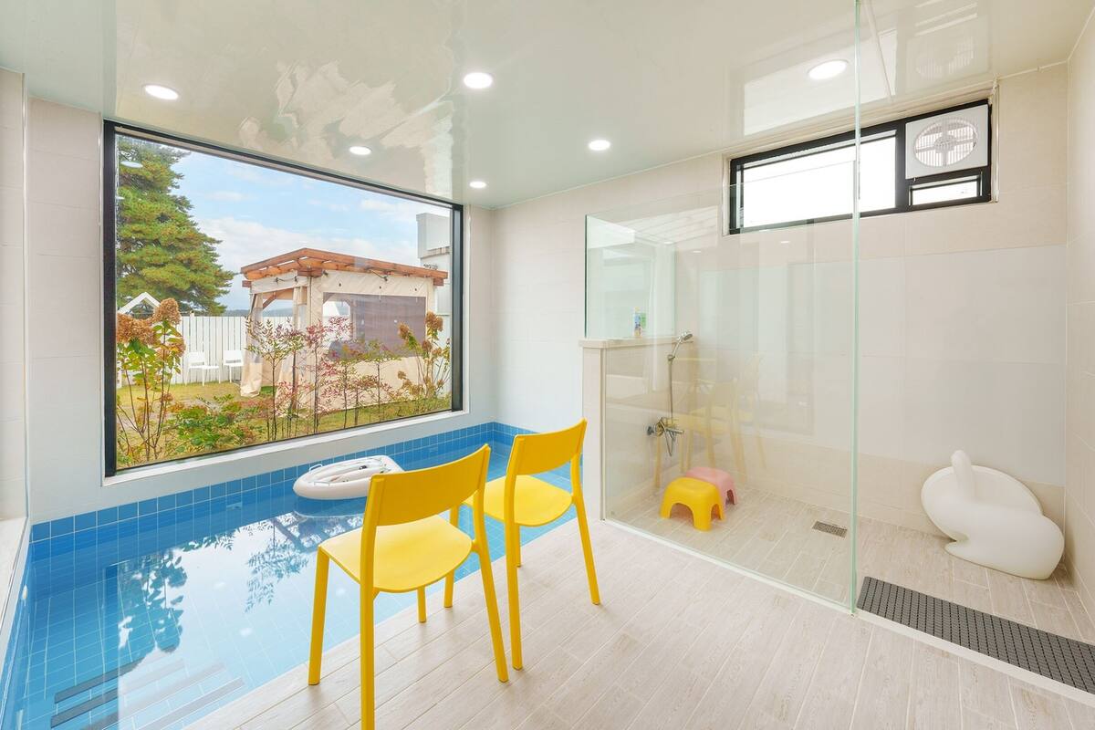 A modern bathroom with a glass shower enclosure, a small pool with yellow chairs, and a view of a house through a large window.