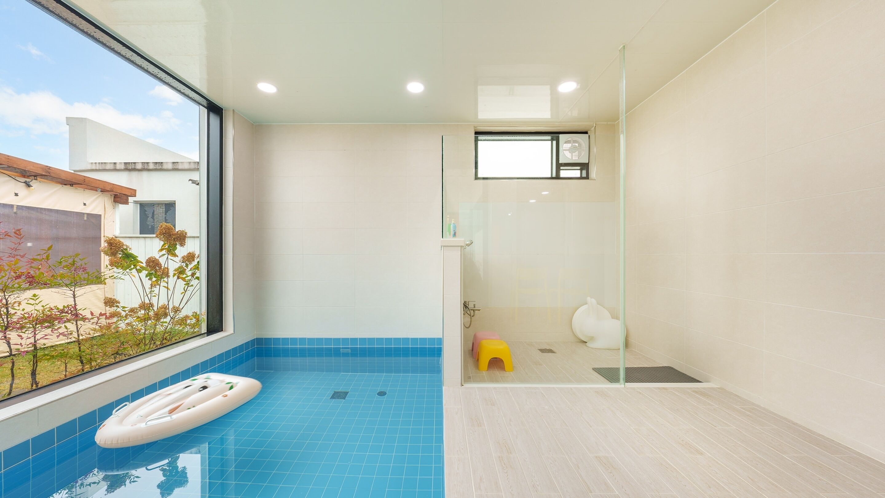 A modern bathroom with a glass shower enclosure, a small yellow stool, and a window overlooking a garden.