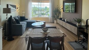 Een moderne woonkamer met een eethoek, grote ramen en een flatscreen televisie aan de muur.