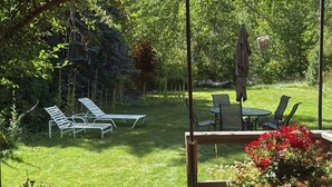 Une terrasse avec une table et des chaises, un parasol rouge et un jardin avec une pelouse et des arbres.