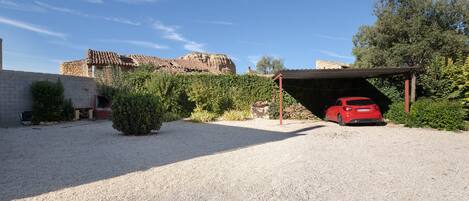 A gravel driveway leading to a red car parked under a covered area, with a stone building and greenery in the background.