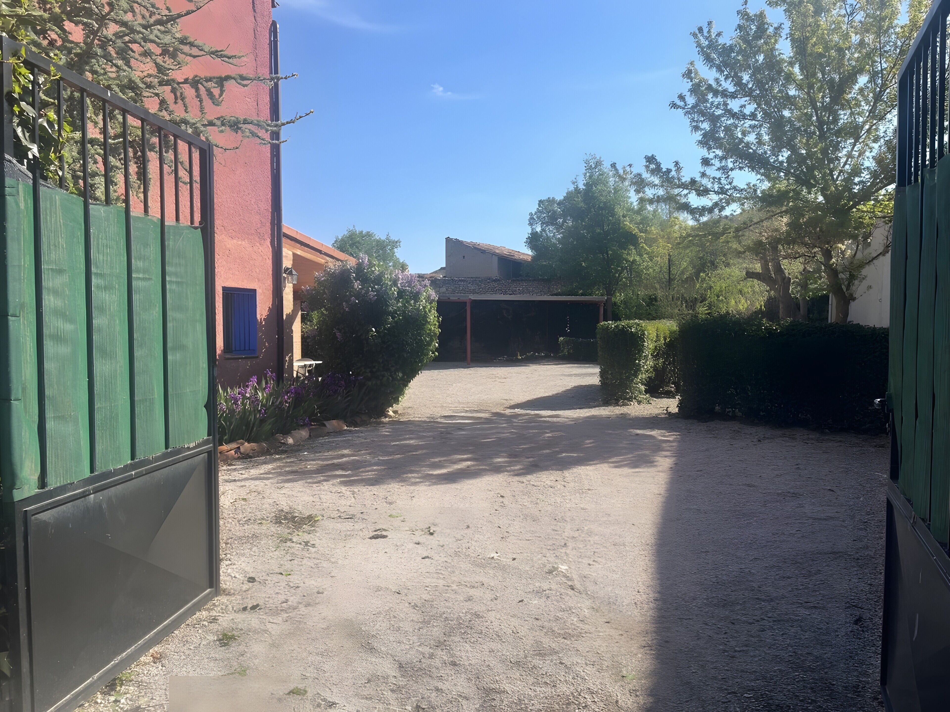 A gravel driveway leading to a building with a covered entrance, surrounded by greenery and a metal gate.
