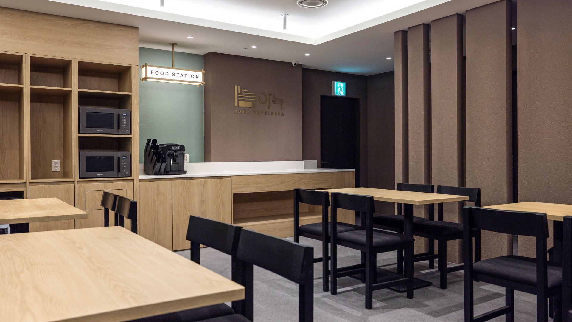 A modern food station area with wooden tables and chairs, a counter with a coffee machine, and a sign indicating the location.