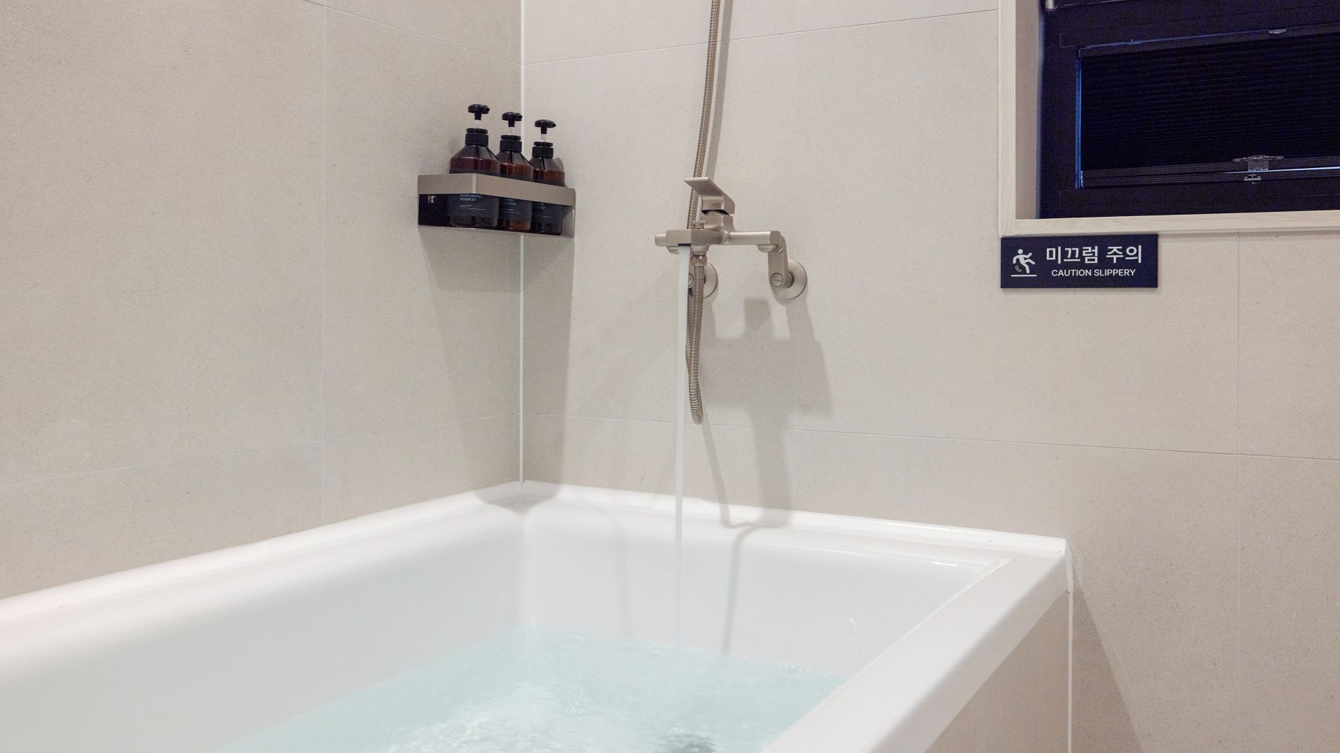 A bathtub with running water, a showerhead, and a shelf with toiletries. A caution sign for slipperiness is visible.