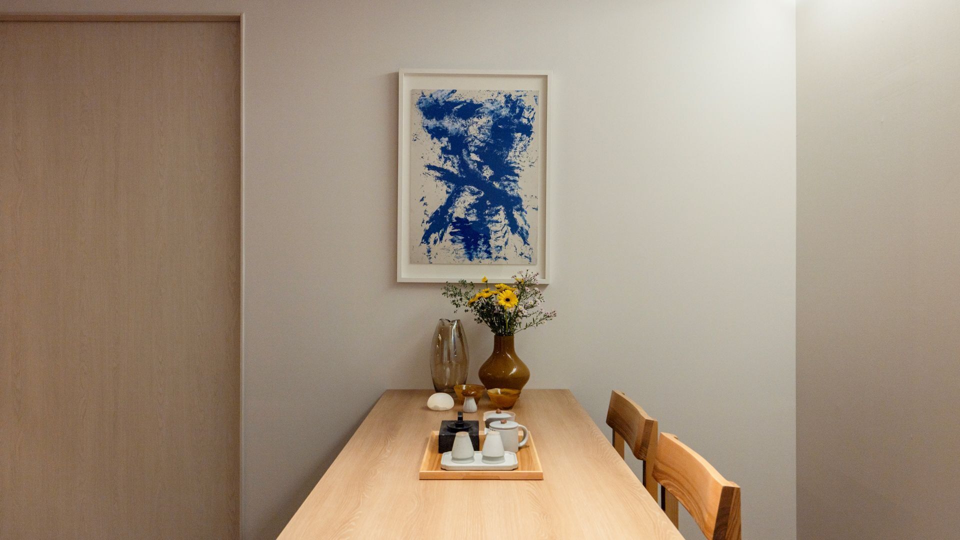 A dining area with a wooden table, a vase with yellow flowers, a framed abstract painting, and a tray with teaware.