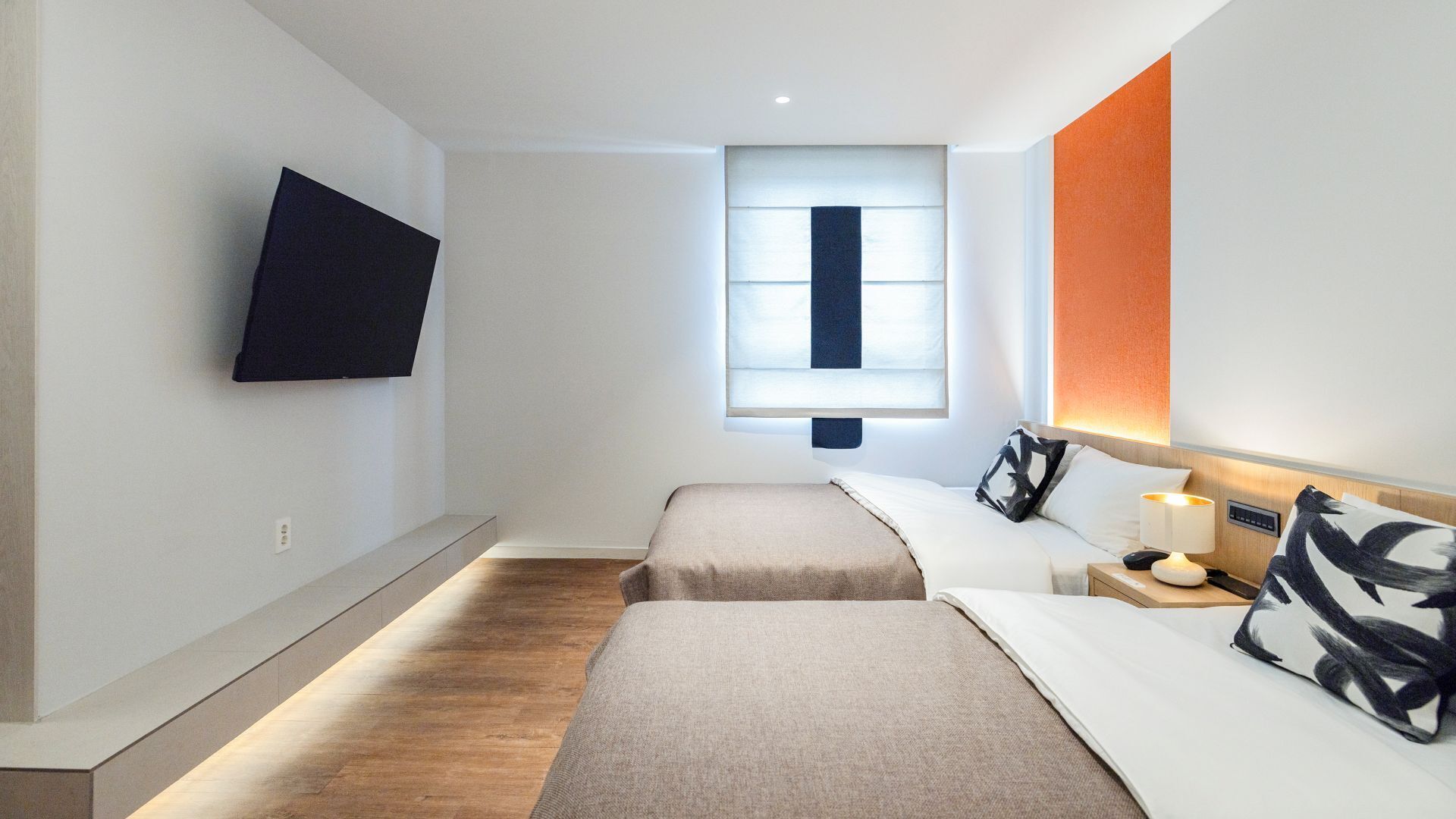 Ein modernes Hotelzimmer mit zwei Betten, einem Wandfernseher, einem Fenster mit Jalousien sowie einem Nachttisch mit Lampe und Telefon.