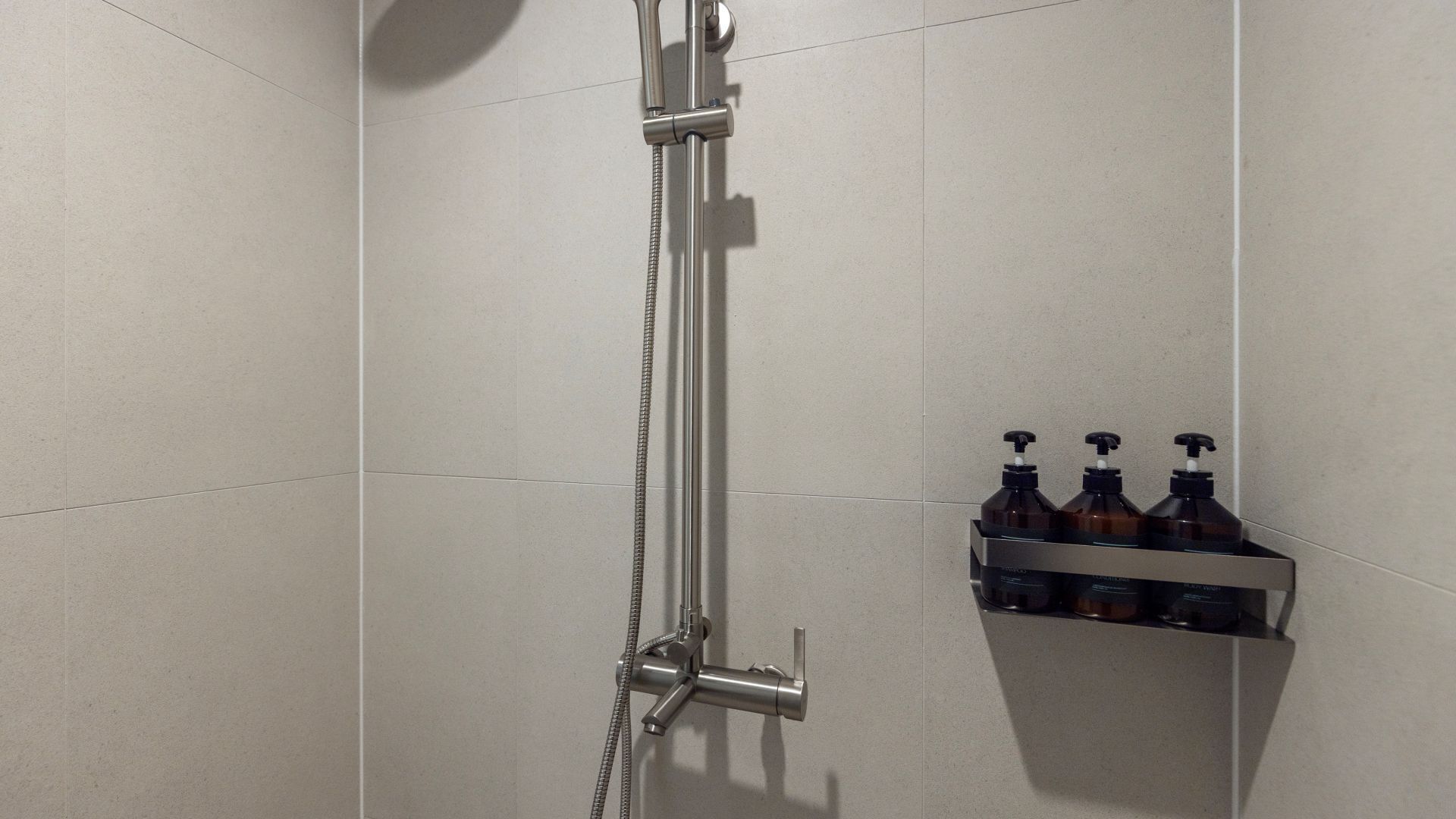 A shower with a handheld showerhead and a wall-mounted faucet. There is a shelf with three bottles of soap.