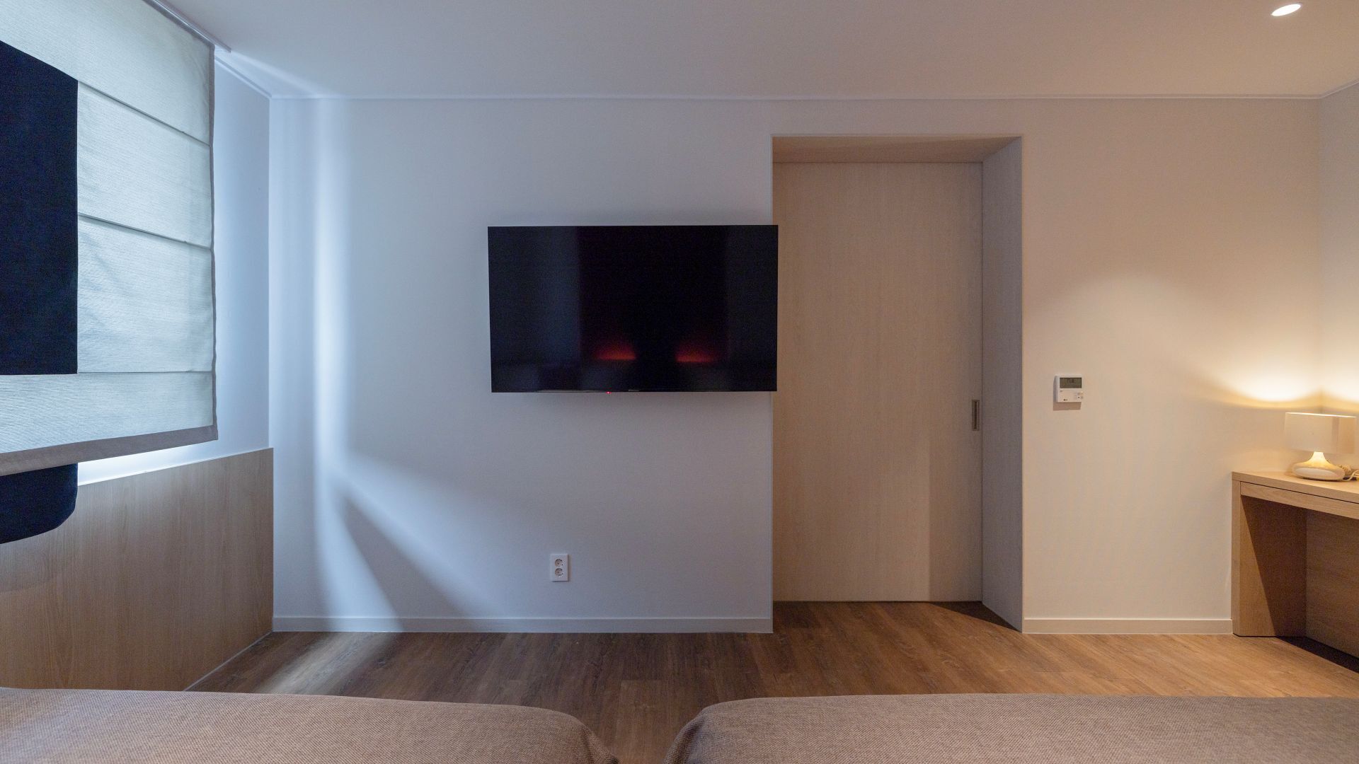 A modern hotel room with a flat-screen TV mounted on the wall, a wooden floor, and a small bedside table with a lamp.