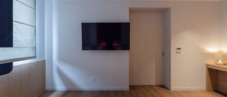 A modern hotel room with a flat-screen TV mounted on the wall, a wooden floor, and a small bedside table with a lamp.
