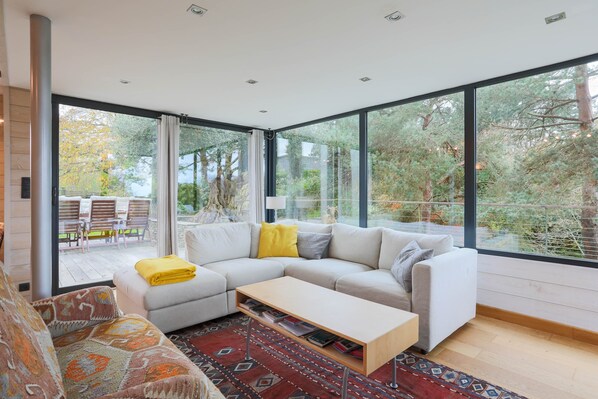 Una sala de estar moderna con un sofá modular, una mesa de centro y amplias ventanas que ofrecen vista a un jardín y un área de estar exterior.