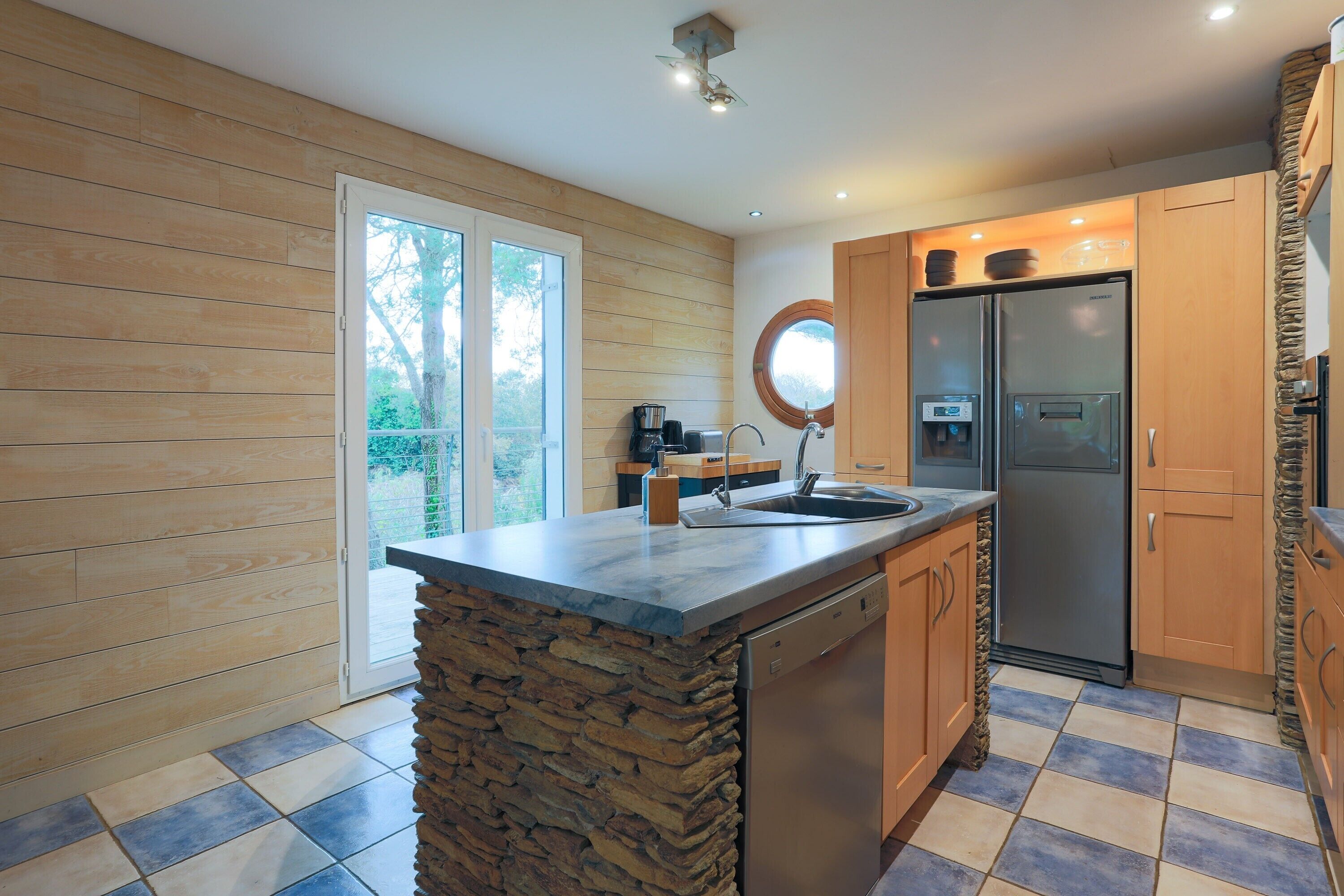 A kitchen with a stone island, wooden cabinets, a stainless steel refrigerator, and a window overlooking greenery.