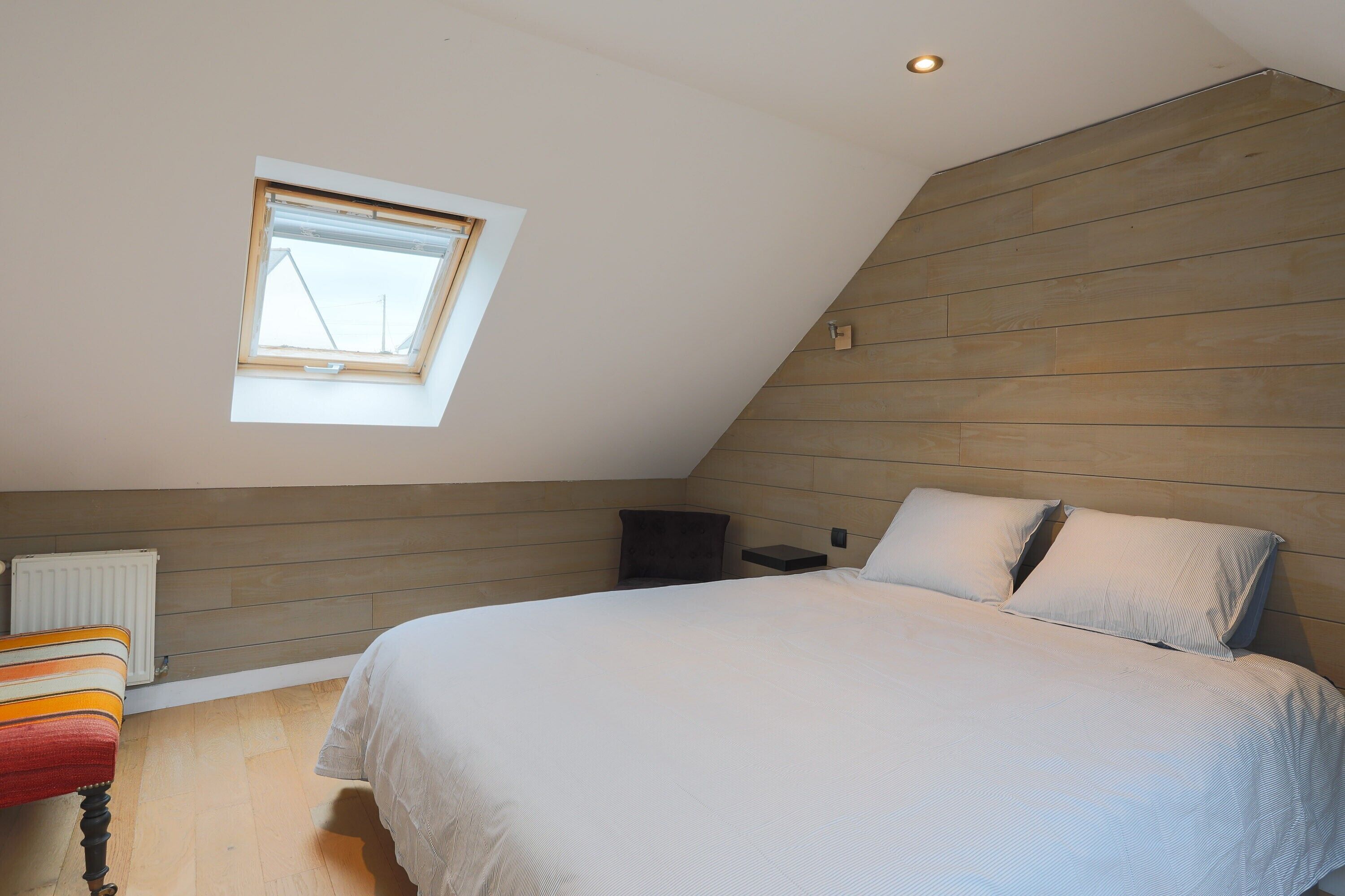 Een slaapkamer met een schuine plafond, dakraam, een bed met kussens, een stoel en een nachtkastje.