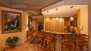 A kitchen with wooden cabinets, a large island with bar stools, a painting of a waterfall, and a potted plant.