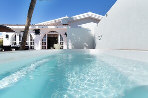 Une piscine à l’eau claire, un bâtiment blanc avec un toit en tuiles et un palmier.