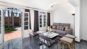 A spacious living area with a sofa, coffee table, and chairs, a glass door leading to a patio with a view of palm trees.