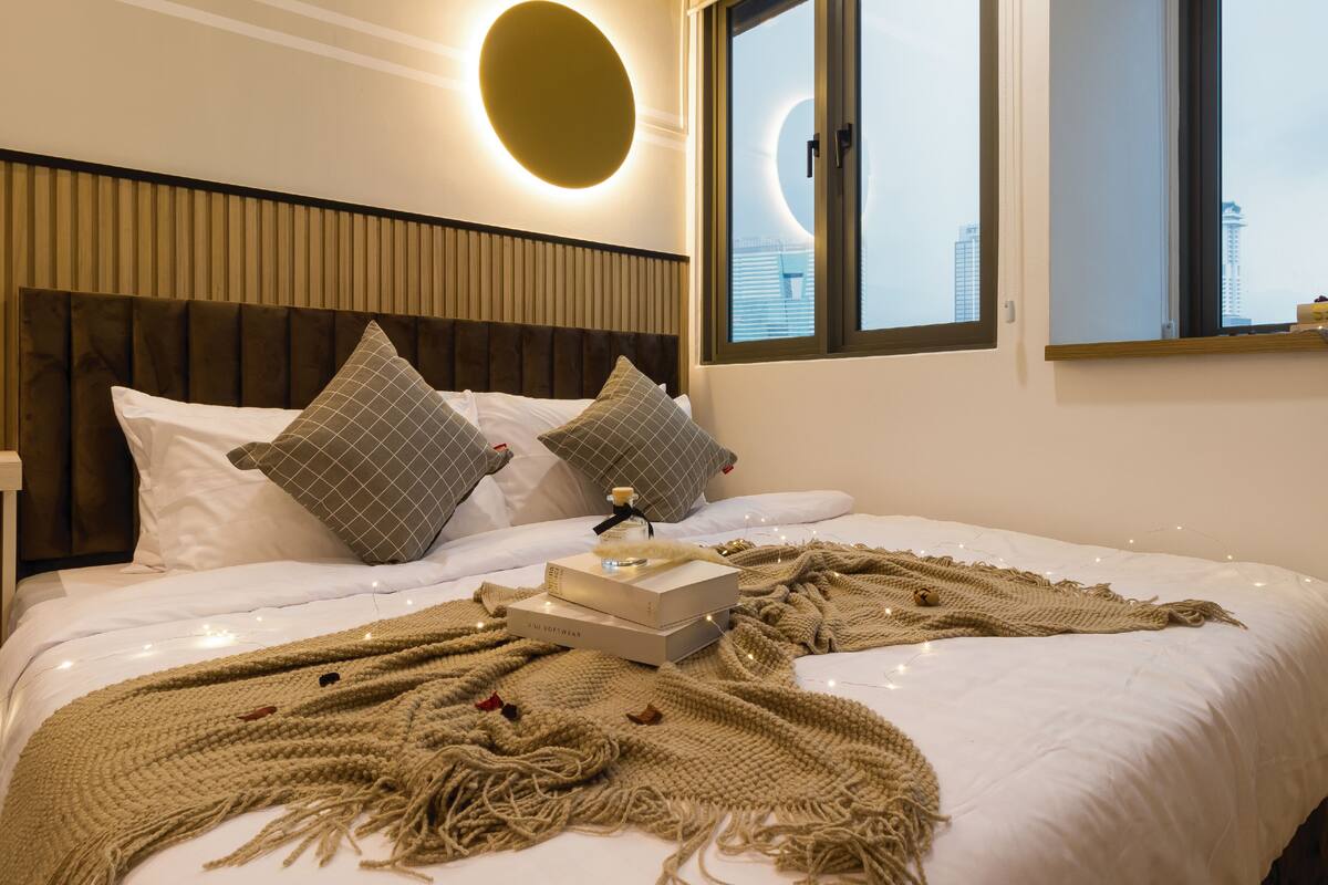 A neatly made bed with a large, circular wall light, two pillows, a blanket, and a book on the bed.