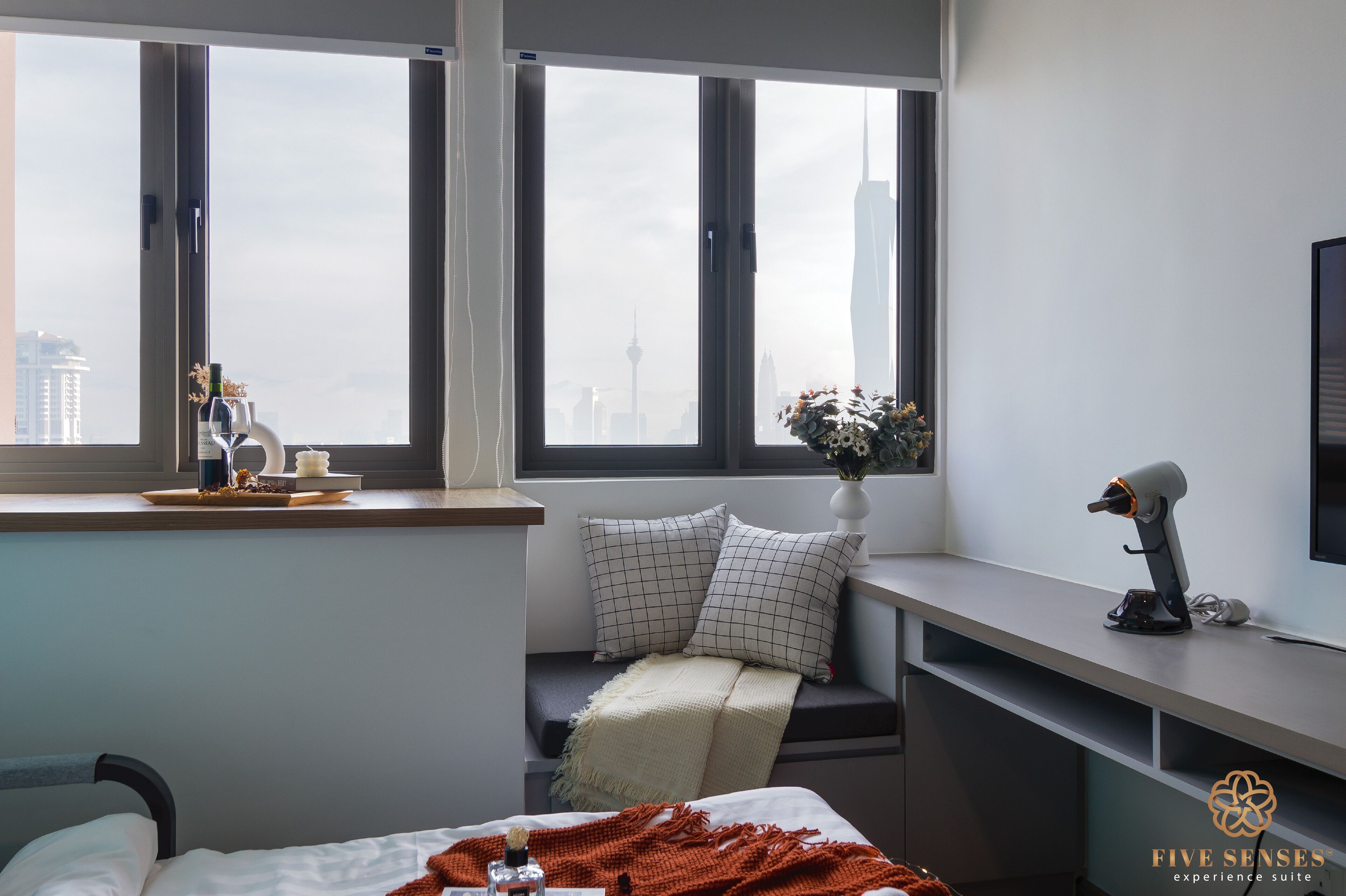 A modern hotel room with a large window, a desk with a lamp, a bed with a blanket, and a view of a city skyline.