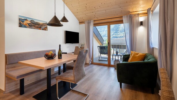 Ein modernes Interieur mit Holzboden, einer Essecke mit Bank und Tisch, einem grünen Sofa und einem großen Fenster mit Bergblick.