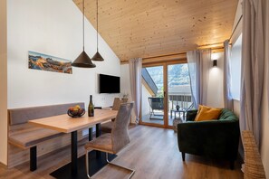 A modern interior with a wooden ceiling, a dining area with a bench and table, a green sofa, and a large window with a view of mountains.