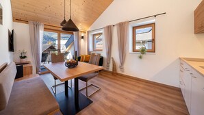 Ein modernes Interieur mit Holzbalkendecke, einem Esstisch mit Stühlen, einer Couch, einer Küchenzeile und großen, mit Vorhängen versehenen Fenstern.
