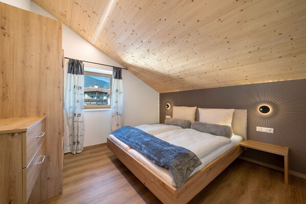 A bedroom with a sloped ceiling, a bed with a headboard, bedside tables, a wardrobe, and a window with curtains.