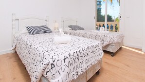 A double bed with alphabet-patterned bedding, a bedside table with a lamp, and a window with a view of palm trees.