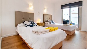 A hotel room with two beds, each with a headboard, bedside tables, and lamps. There is a balcony with a view.