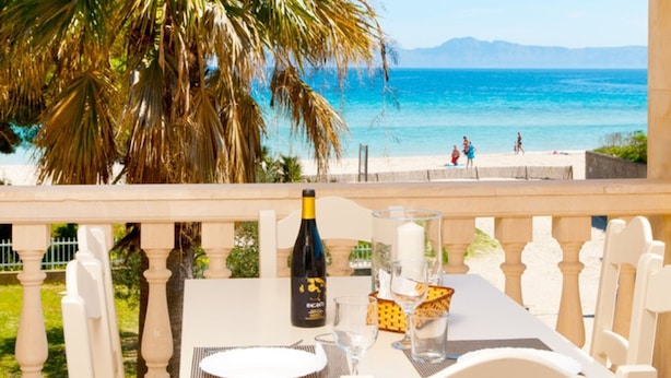 Ein Tisch mit einer Flasche, Gläsern und einem Teller auf einem Balkon mit Blick auf einen Strand, im Hintergrund Personen.