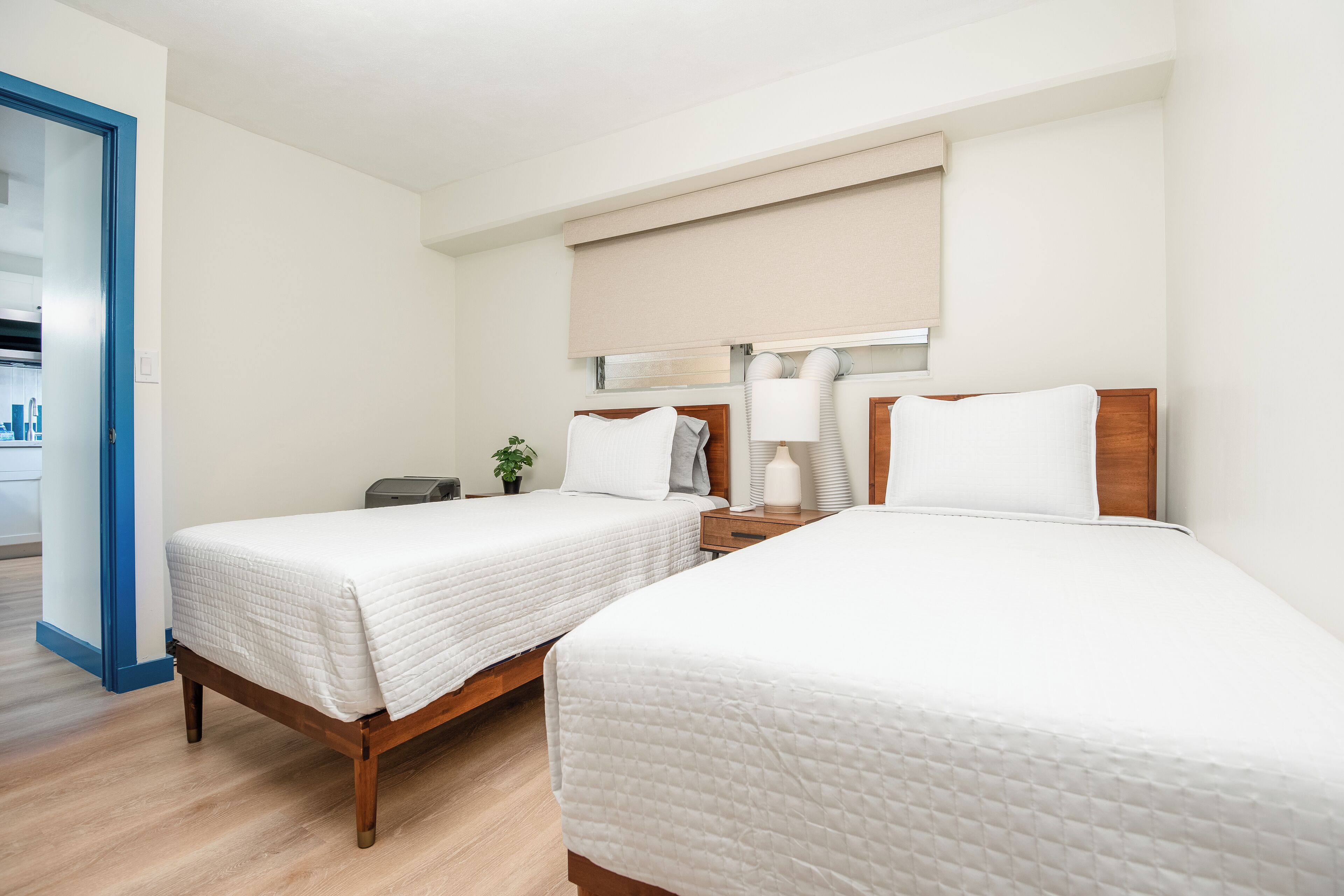 Une chambre d’hôtel avec deux lits, une table de chevet avec une lampe, une petite plante et une fenêtre avec un store beige.