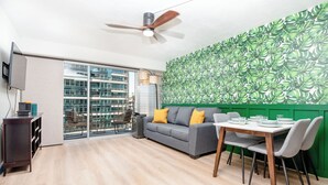 A modern living room with a grey sofa, wooden dining table, and chairs. There is a ceiling fan, a TV mounted on the wall, and a green leafy wallpaper. - Royal Aloha by Tropical Escapes Realty (Honolulu)